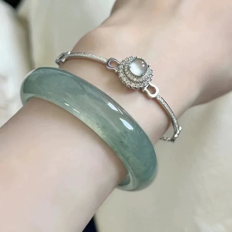 A smooth ice oil green jadeite bangle and a decorative silver-toned bracelet on a woman's wrist.